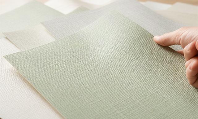 淡い緑色と細かな織り柄の壁紙サンプルを室内の光で見比べている様子